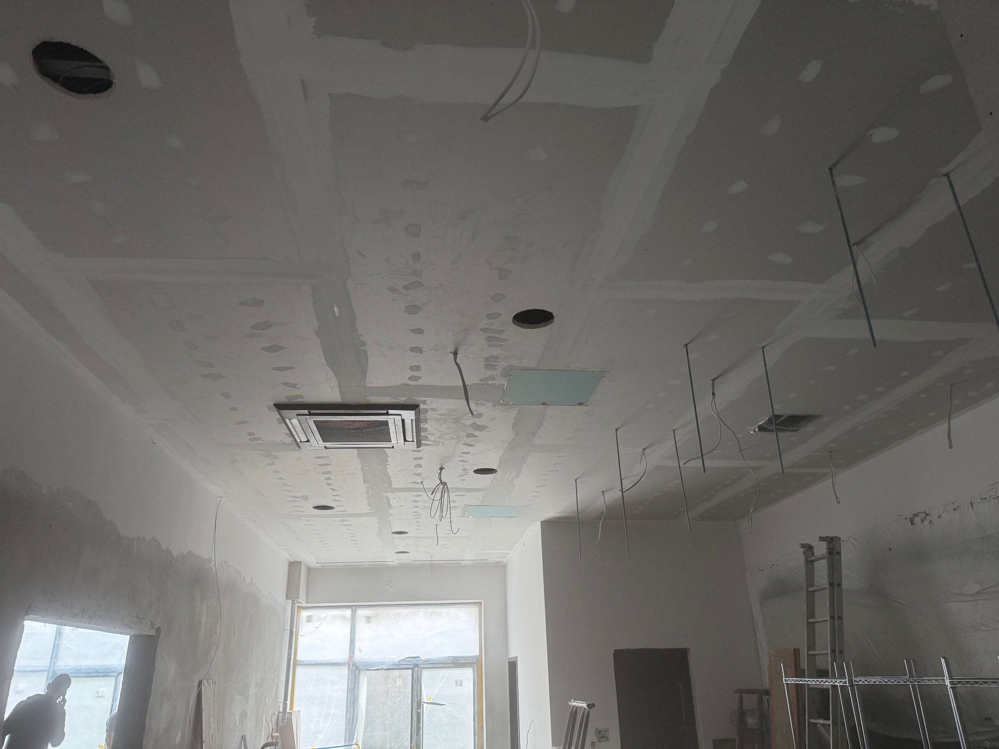 Sufit z płyt karton-gips na metalowym stelażu w trakcie montażu, widoczne otwory na oświetlenie i instalacje. Ściany w trakcie wykończenia, drabina w tle.