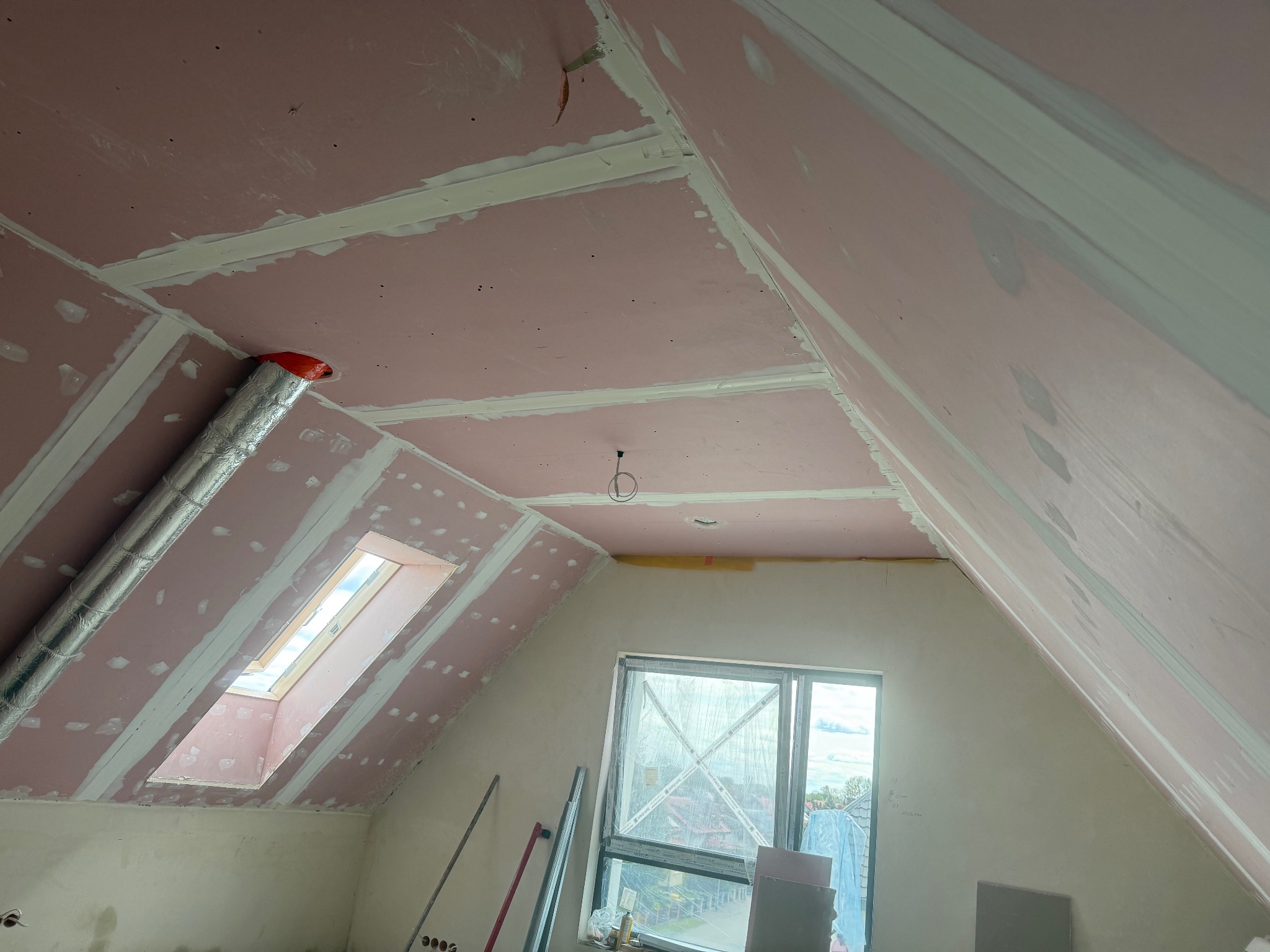 Poddasze z oknem dachowym w trakcie wykańczania: różowe płyty karton-gips na skosach, szpachlowane łączenia. Widoczne narzędzia i fragment okna.