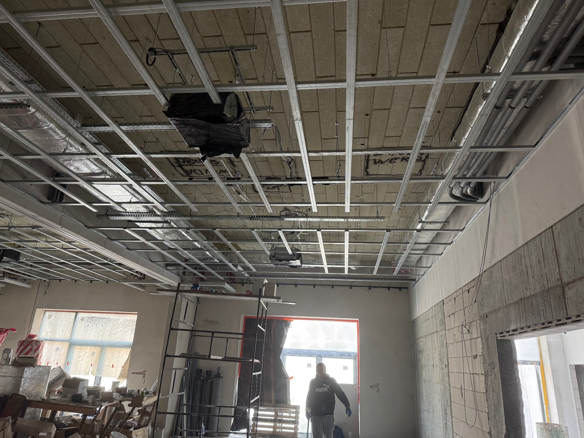 Montaż stelaża sufitowego w Rymań. Widoczne profile metalowe, izolacja i instalacje. W tle okno i pracownik w bluzie. Surowy stan wykończenia wnętrza.