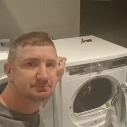 Mężczyzna opiera się o białą suszarkę Bosch z otwartymi drzwiczkami, na górze leży scyzoryk, w tle kafelki i półka.