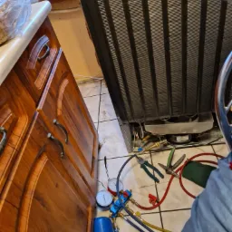 Tył lodówki z widocznym agregatem, narzędzia serwisowe rozłożone na podłodze, manometry, węże, szczypce, obok szafka kuchenna.