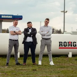 Trzech mężczyzn pozuje na tle zbiornika z logo ROMGAZ i stacji paliw w tle.