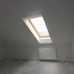 Poddasze po remoncie z oknem dachowym, grzejnikiem i świeżo pomalowanymi ścianami na biało, widoczna surowa podłoga i drewniana belka konstrukcyjna.