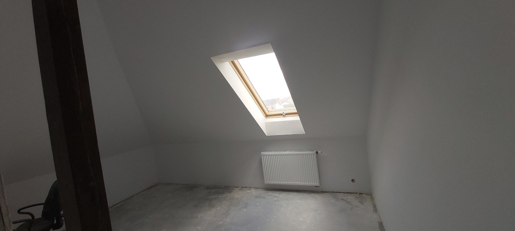 Poddasze po remoncie z oknem dachowym, grzejnikiem i świeżo pomalowanymi ścianami na biało, widoczna surowa podłoga i drewniana belka konstrukcyjna.