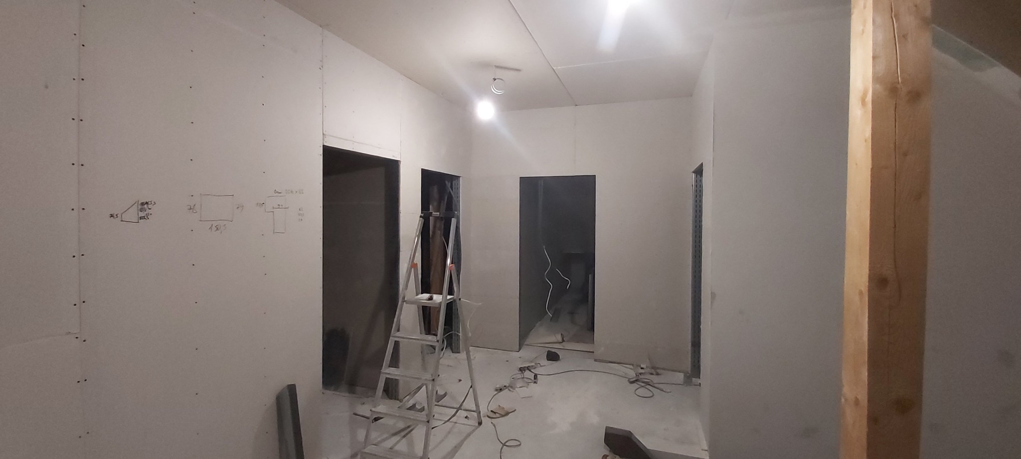 Wnętrze w trakcie wykańczania z płyt kartonowo-gipsowych, widoczne szkice wymiarów na ścianie, drabina aluminiowa i drewniany słup konstrukcyjny.