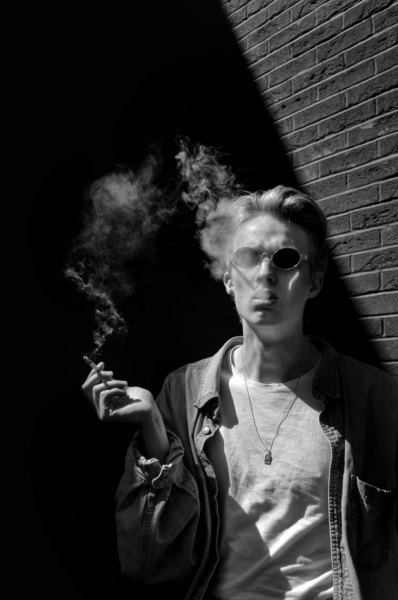 Czarno-białe zdjęcie młodego mężczyzny w okularach przeciwsłonecznych, opierającego się o ceglaną ścianę, z dymem papierosowym unoszącym się w powietrzu.