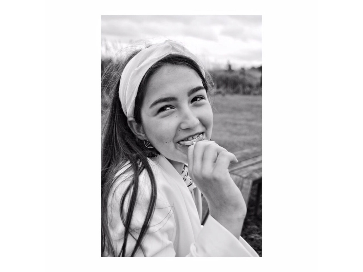 Portret uśmiechniętej nastolatki w plenerze, czarno-białe zdjęcie z opaską na włosach i delikatną biżuterią.