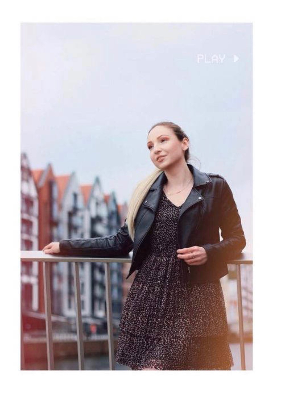 Kobieta w skórzanej kurtce i wzorzystej sukience, oparta o barierkę z widokiem na budynki w tle, stylizowane ujęcie z efektem retro 'PLAY'