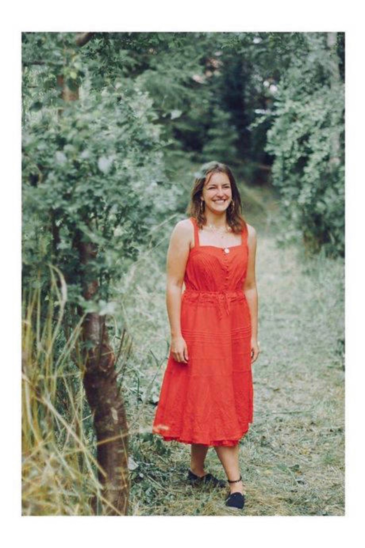 Uśmiechnięta kobieta w czerwonej sukience stoi na leśnej ścieżce, z delikatnym oświetleniem i naturalnym tłem.