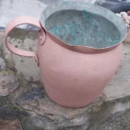 Miedziany dzbanek z uchwytem, częściowo pokryty patyną, stojący na kamiennej powierzchni, z fragmentem oczka wodnego w tle.