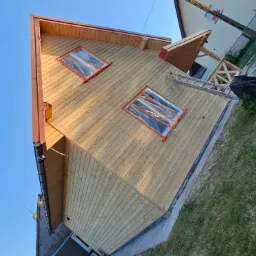 Drewniany dom w trakcie renowacji elewacji, okna zabezpieczone folią, widoczna drabina i elementy drewnianej konstrukcji werandy, jasne drewno elewacyjne.