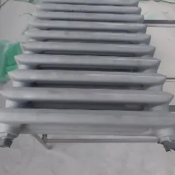 Stos grzejników żeliwnych po obróbce, pokrytych świeżą, jednolitą warstwą szarej farby, ustawionych na metalowym stelażu w pomieszczeniu z zabezpieczoną folią ścianą.