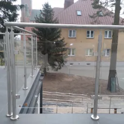 Balkon z nowoczesną szklaną balustradą i elementami ze stali nierdzewnej, widok na budynek mieszkalny i kościół w tle.