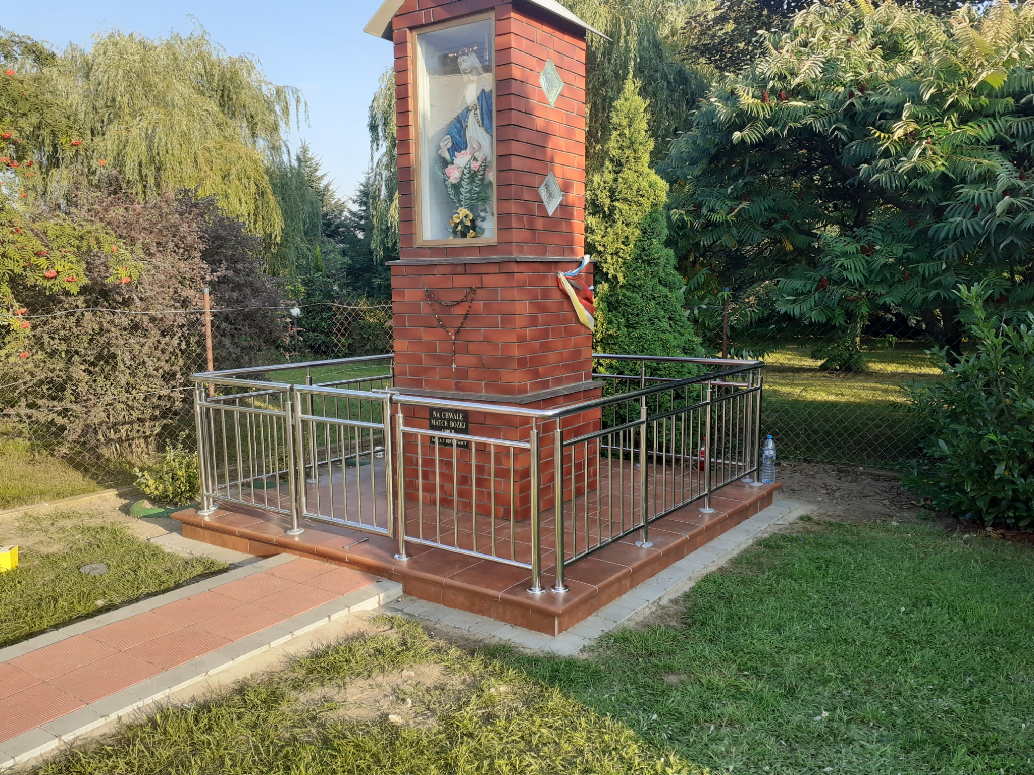 Kapliczka z cegły otoczona niską, metalową barierką, z widocznym napisem i elementami dekoracyjnymi, umieszczona w ogrodzie.