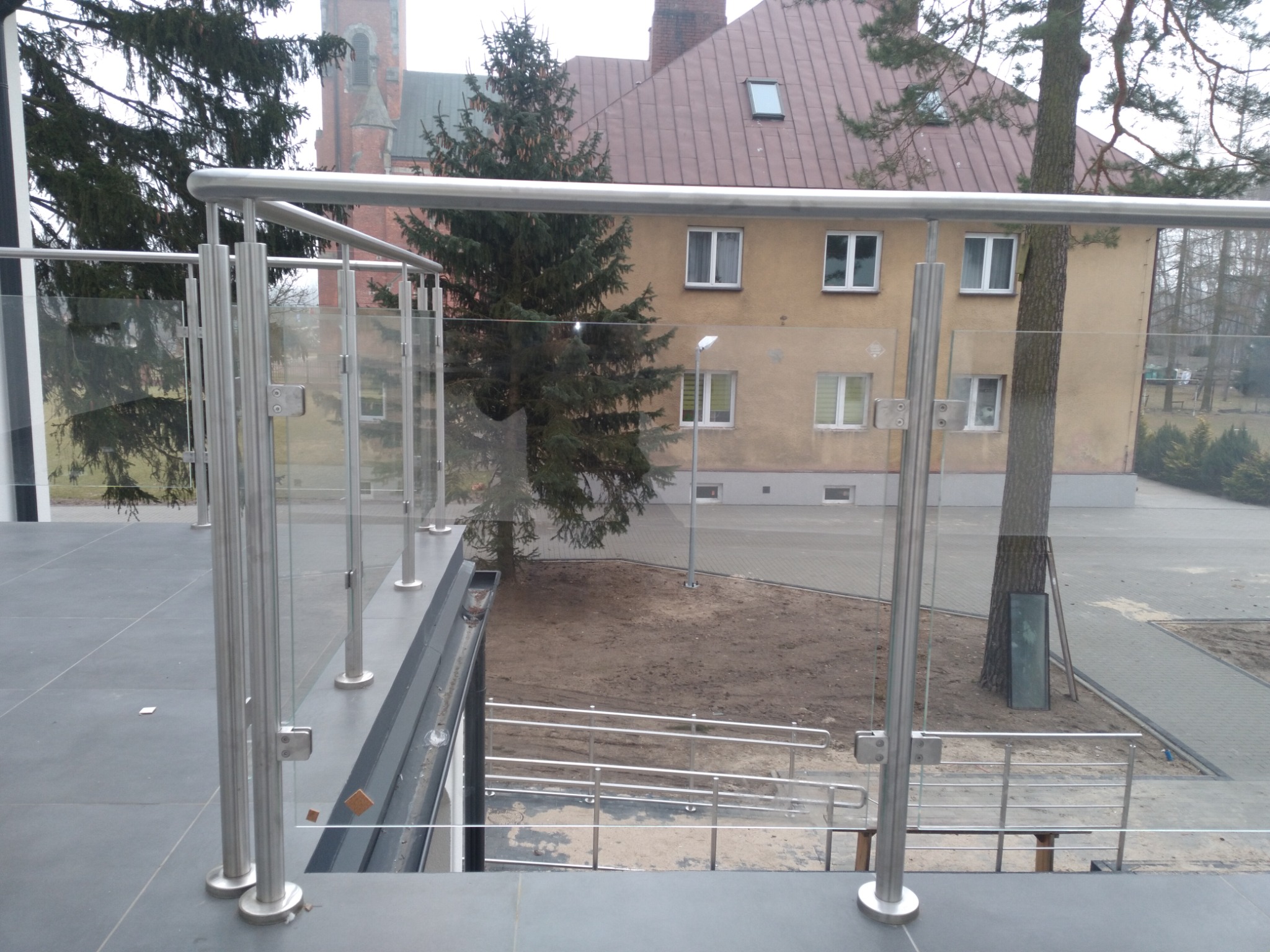 Balkon z nowoczesną szklaną balustradą i elementami ze stali nierdzewnej, widok na budynek mieszkalny i kościół w tle.