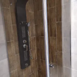 Kabina prysznicowa z czarnym panelem natryskowym z hydromasażem, wykończona płytkami imitującymi drewno w odcieniach brązu, z odpływem liniowym w podłodze.