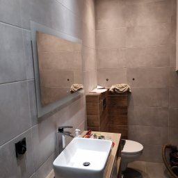 RAMAK Robert Mieszczanin - Nowoczesna łazienka w trakcie remontu: biała umywalka nablatowa, bateria, lustro z podświetleniem LED, szare płytki wielkoformatowe, drewniana obudowa stelaża WC, narzędzia na blacie, brakująca...