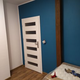 RAMAK Robert Mieszczanin - Pomalowane na niebiesko ściany w pokoju z drewnianą belką, białe drzwi z przeszkleniami i podłoga z paneli, materac w folii i zabawka na podłodze.