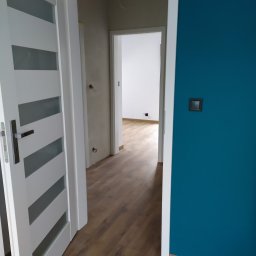 RAMAK Robert Mieszczanin - Nowoczesne wnętrze mieszkania po remoncie: turkusowa ściana, białe drzwi z przeszkleniami i podłoga z paneli imitujących drewno, widok na kolejne pomieszczenie z surową ścianą i wystającymi kablami.