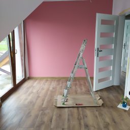 RAMAK Robert Mieszczanin - Pokój na poddaszu z drewnianą podłogą, ściana pomalowana na różowo, aluminiowa drabina na kartonie, widoczne narzędzie malarskie i roślina w niebieskiej doniczce.