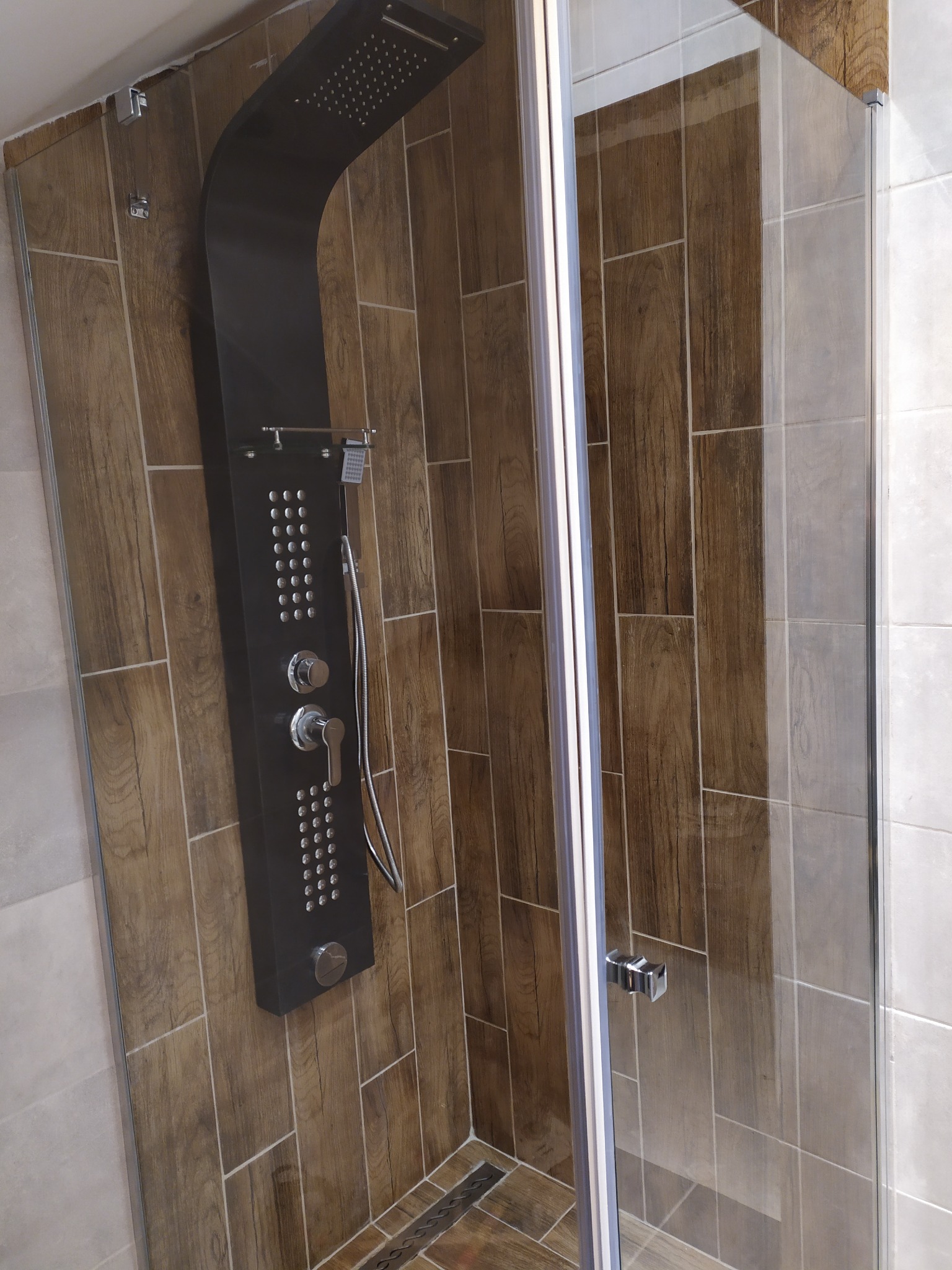 Kabina prysznicowa z czarnym panelem natryskowym z hydromasażem, wykończona płytkami imitującymi drewno w odcieniach brązu, z odpływem liniowym w podłodze.