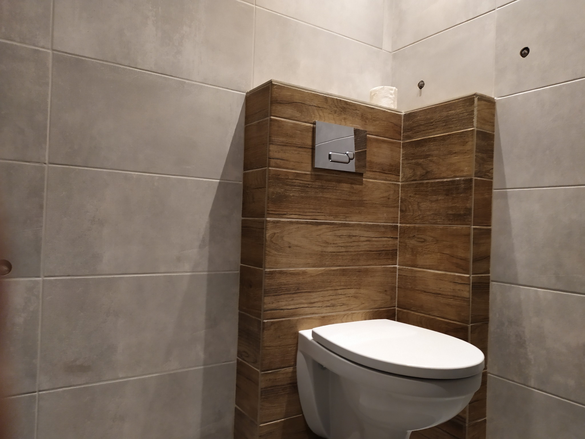 Nowoczesna toaleta podwieszana z drewnopodobną obudową stelaża i szarymi, wielkoformatowymi płytkami na ścianach, widoczne metalowe przyciski spłukiwania.
