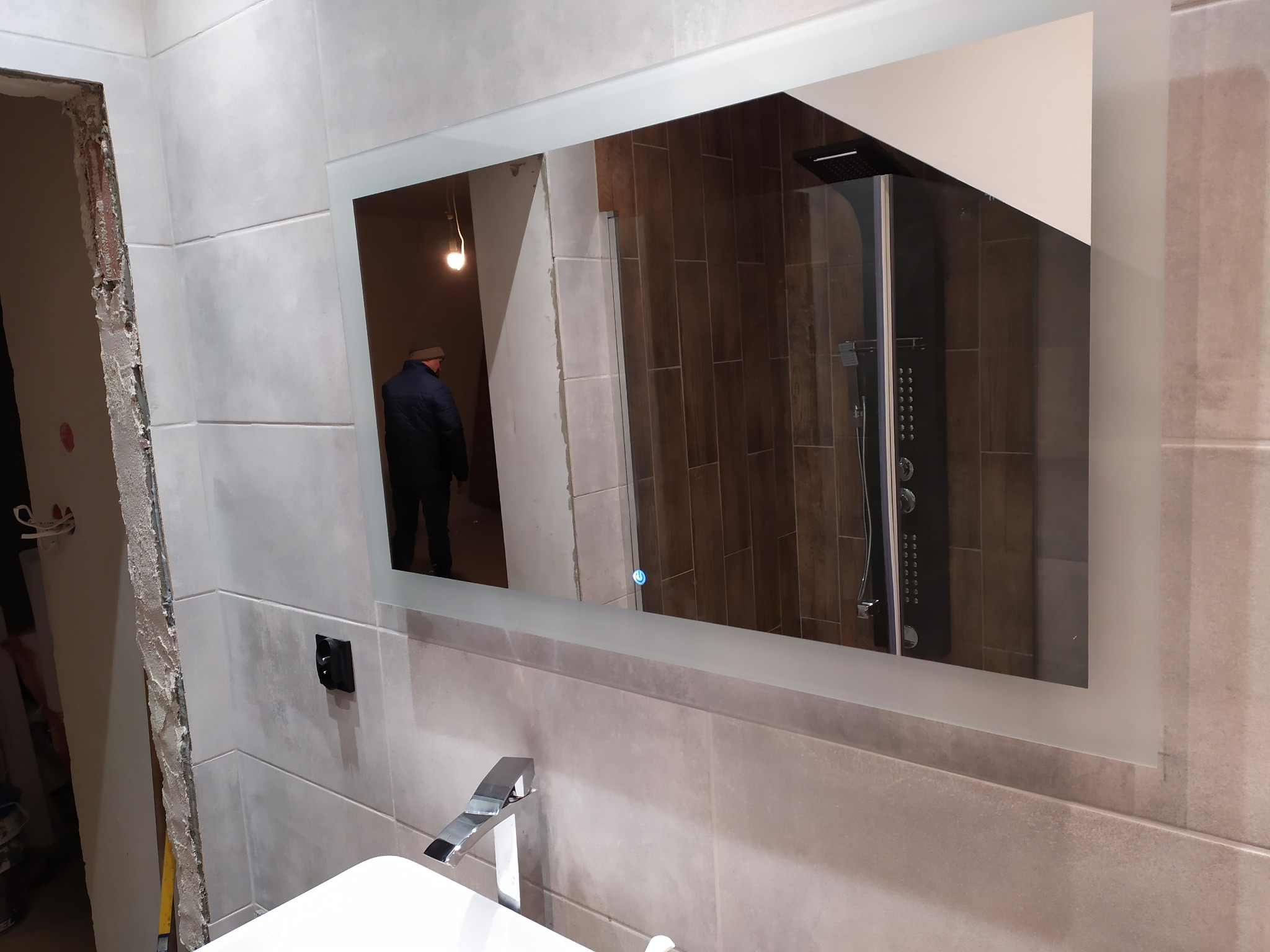 Łazienka w trakcie remontu, widoczne szare płytki, biała umywalka z chromowaną baterią, lustro z podświetleniem LED, w odbiciu kabina prysznicowa i fragment pomieszczenia z instalacją elektryczną.
