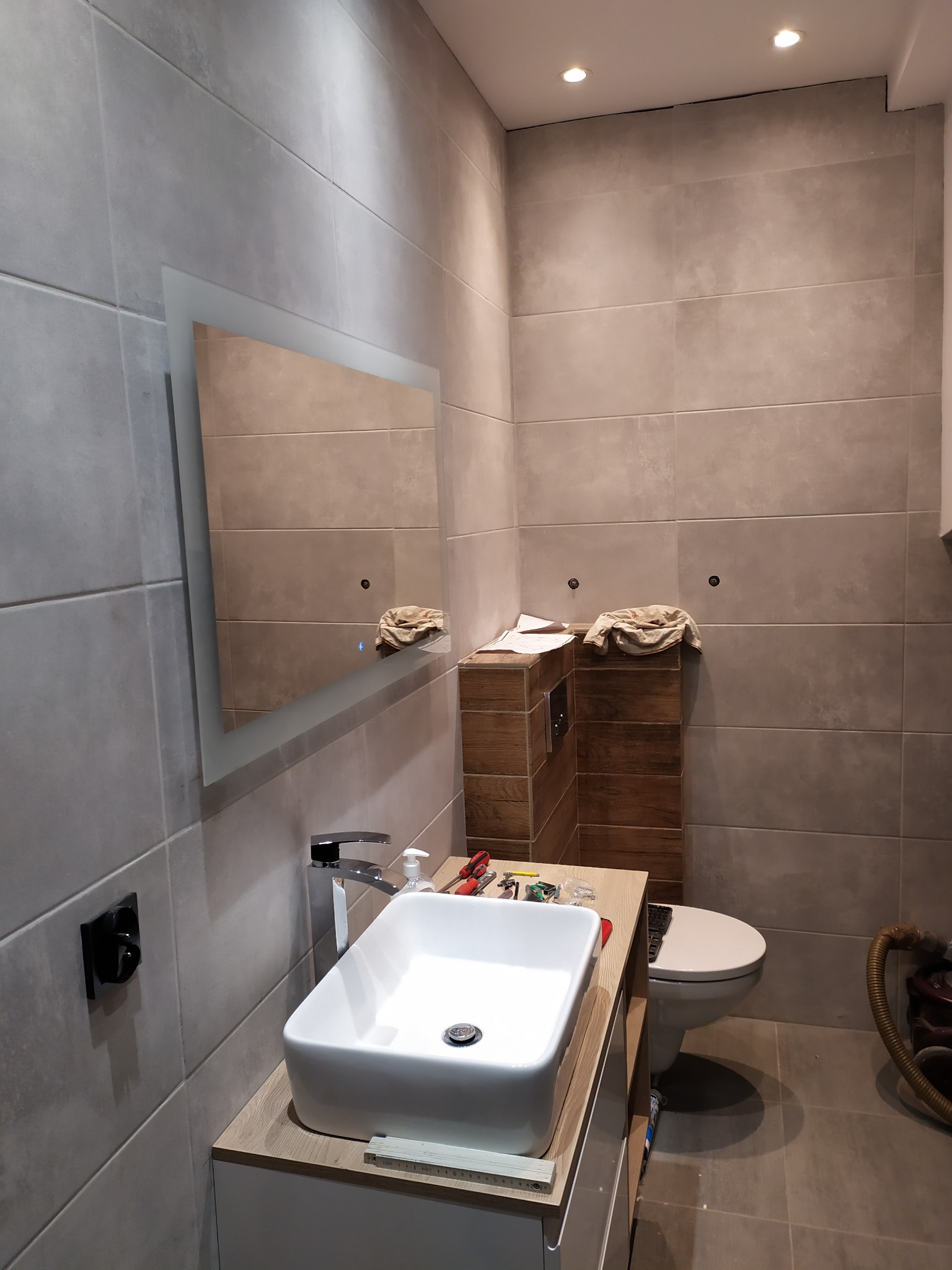 Nowoczesna łazienka w trakcie remontu: biała umywalka nablatowa, bateria, lustro z podświetleniem LED, szare płytki wielkoformatowe, drewniana obudowa stelaża WC, narzędzia na blacie, brakująca...