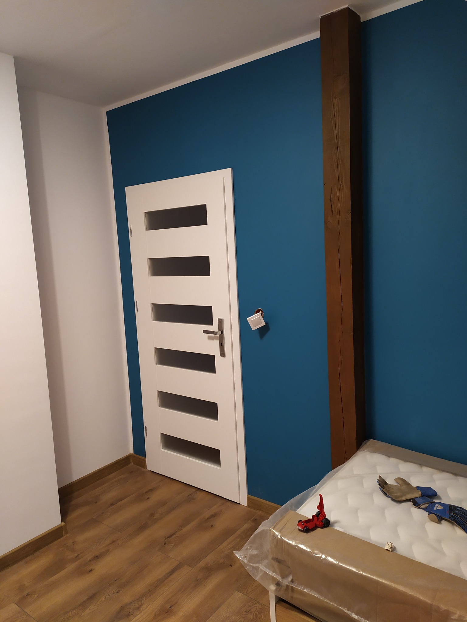 Pomalowane na niebiesko ściany w pokoju z drewnianą belką, białe drzwi z przeszkleniami i podłoga z paneli, materac w folii i zabawka na podłodze.