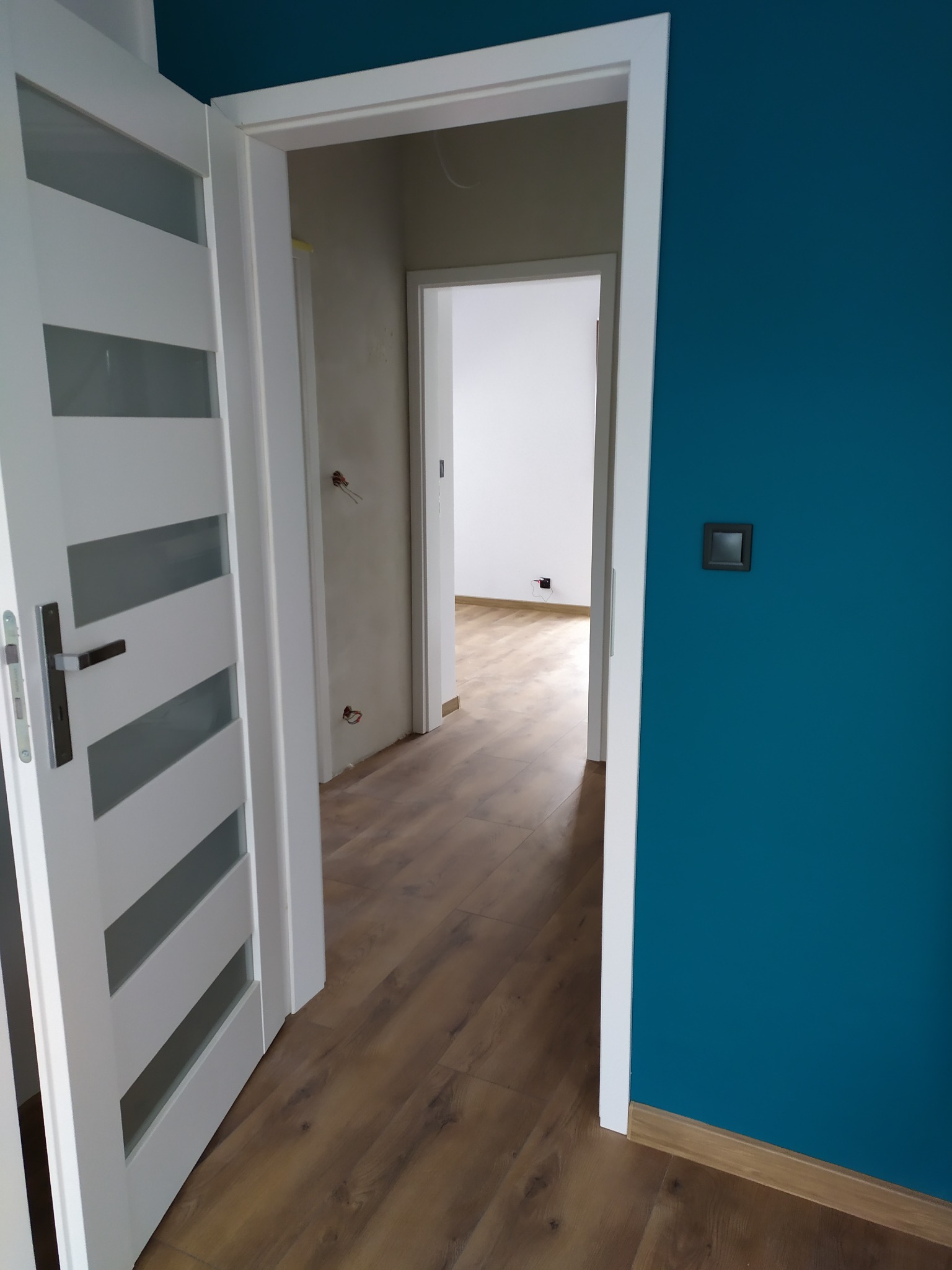 Nowoczesne wnętrze mieszkania po remoncie: turkusowa ściana, białe drzwi z przeszkleniami i podłoga z paneli imitujących drewno, widok na kolejne pomieszczenie z surową ścianą i wystającymi kablami.