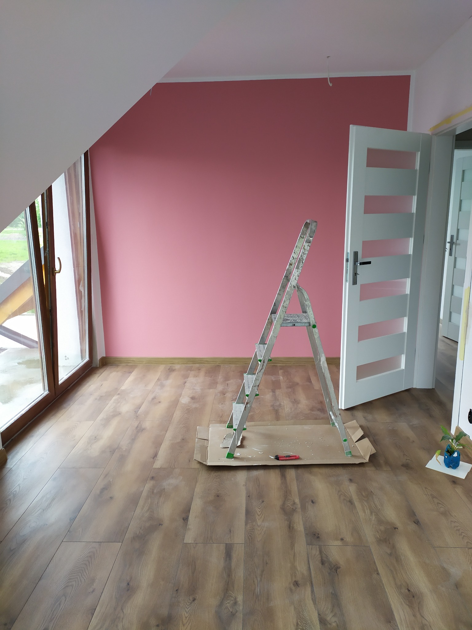 Pokój na poddaszu z drewnianą podłogą, ściana pomalowana na różowo, aluminiowa drabina na kartonie, widoczne narzędzie malarskie i roślina w niebieskiej doniczce.