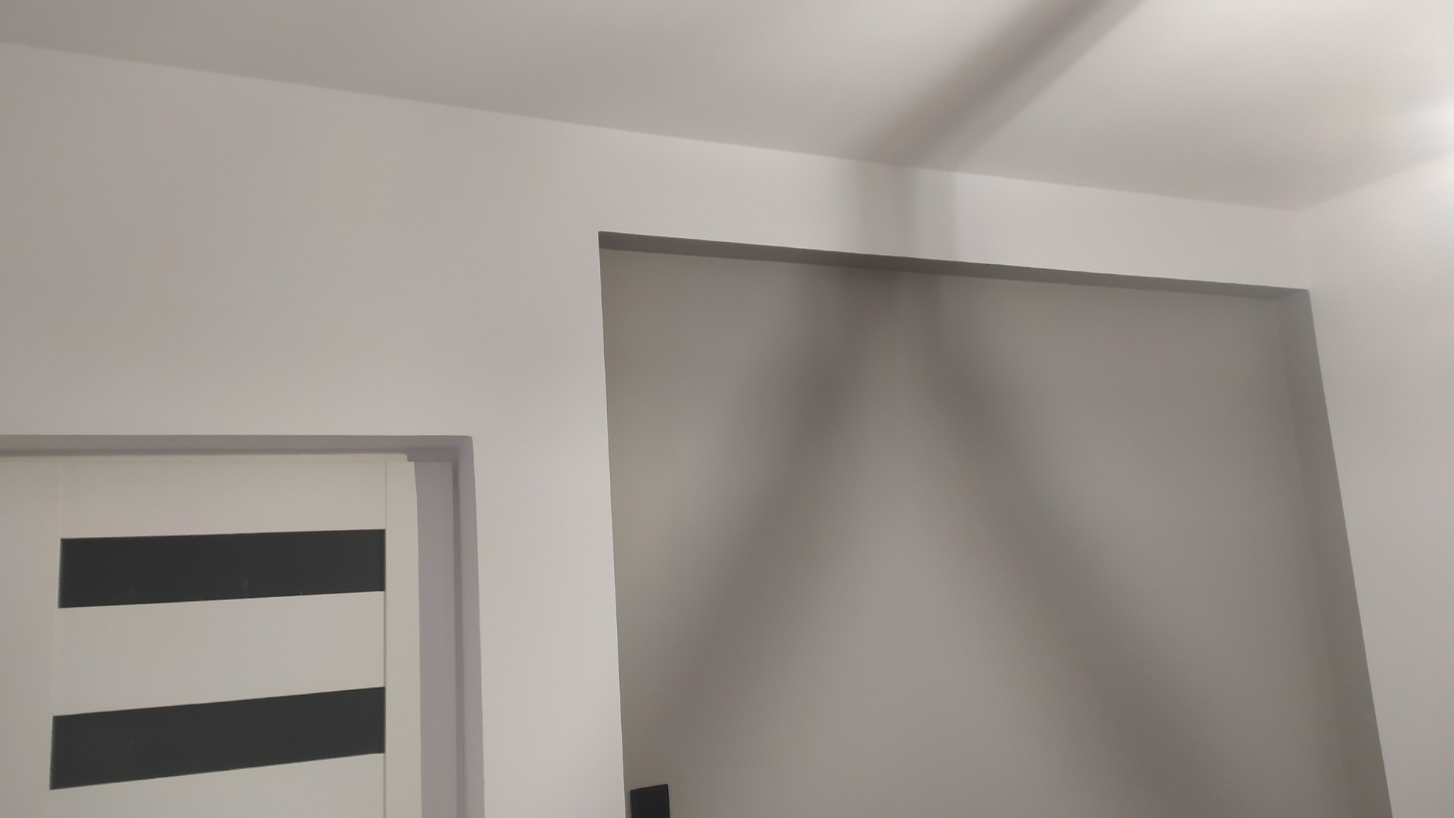 Świeżo pomalowane na biało ściany z widocznym otworem drzwiowym bez ościeżnicy i fragmentem drzwi z poziomymi czarnymi pasami, ukazujące minimalistyczny design wnętrza.