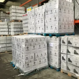 Palety z zapakowanymi kartonami w magazynie, widoczne etykiety z nazwami wódek 'Adam Mickiewicz' oraz 'Ogiński' z motywem fortepianu.