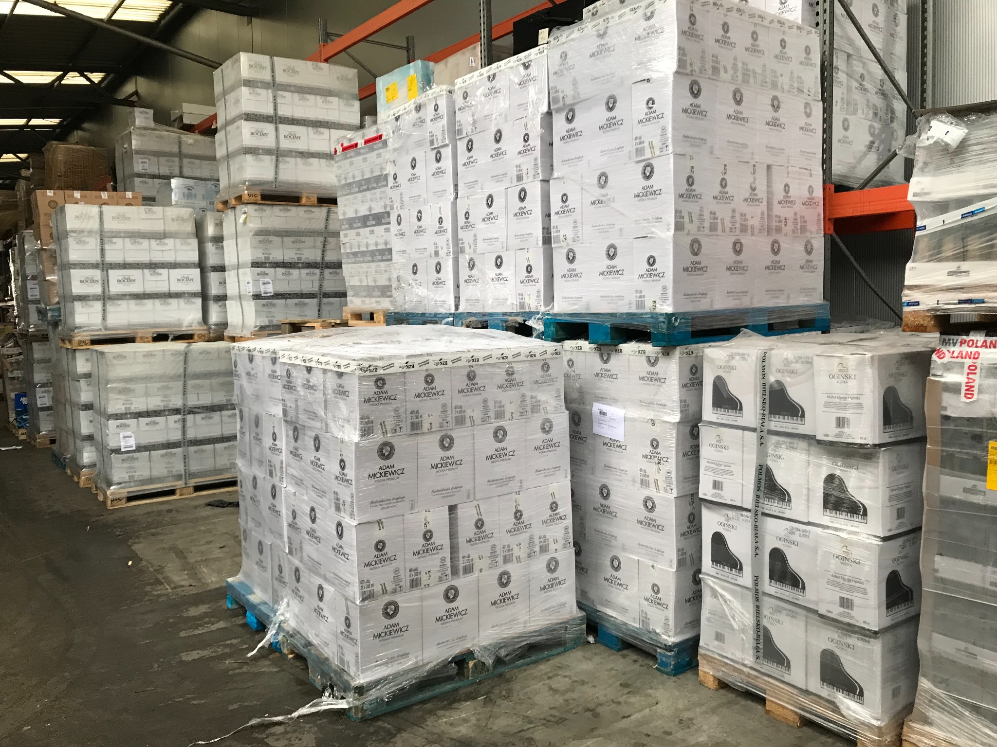 Palety z zapakowanymi kartonami w magazynie, widoczne etykiety z nazwami wódek 'Adam Mickiewicz' oraz 'Ogiński' z motywem fortepianu.