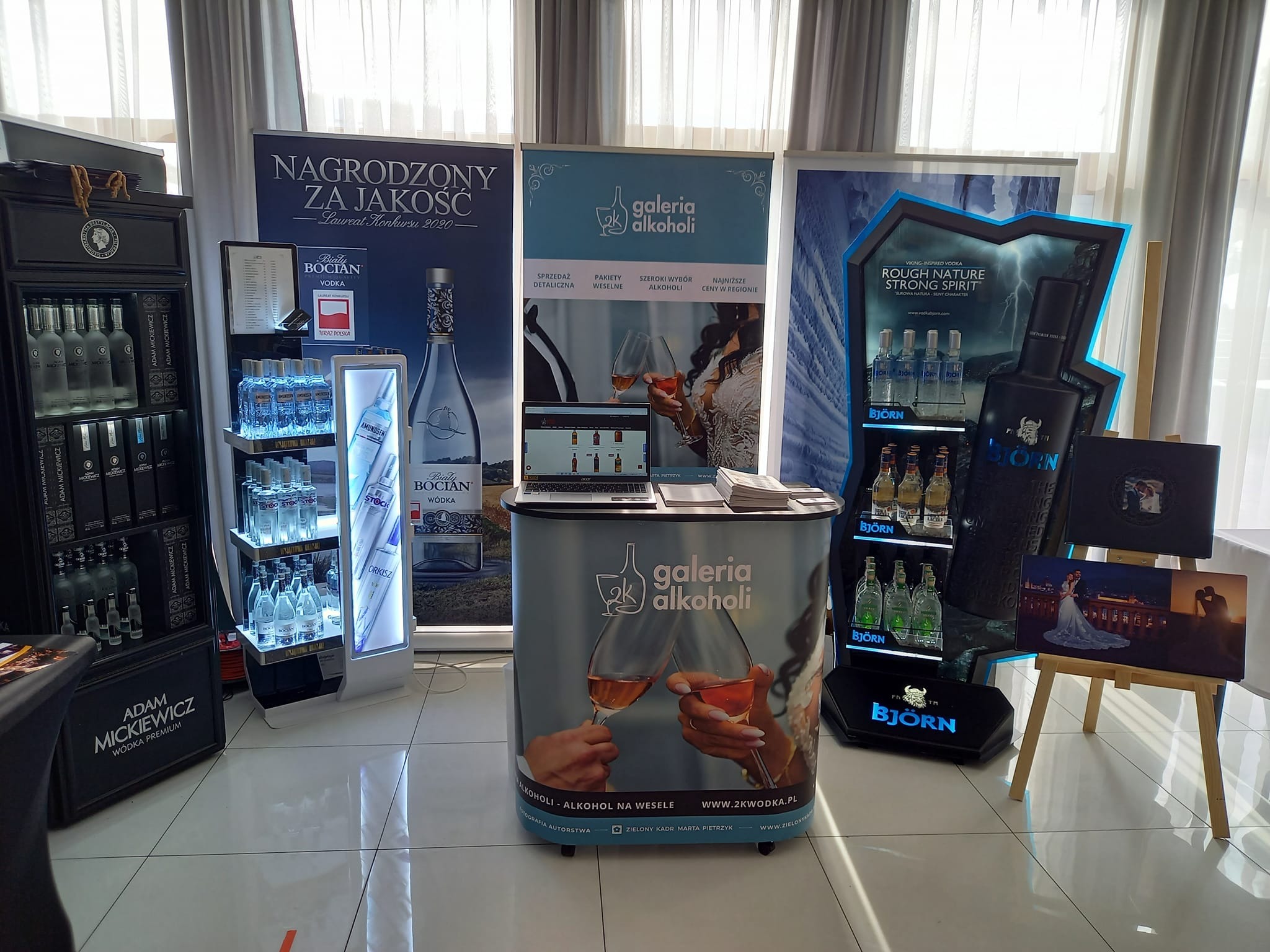 Ekspozycja różnych marek alkoholi na stoisku targowym, w tym wódki Adam Mickiewicz, Biały Bocian i Bjorn, z elementami dekoracyjnymi i reklamowymi, w tle zasłony okienne.