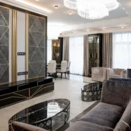 Elegancki salon z marmurową ścianą z geometrycznym wzorem w kolorze złota, czarnym stolikiem RTV i szklanymi stolikami kawowymi na białej podłodze.