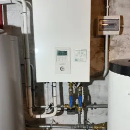 Zamontowana, biała pompa ciepła Panasonic Aquarea Smart Cloud, widoczne rury instalacyjne, zawory z niebieskimi uchwytami, manometr i panel sterowania, na tle surowej ściany z betonu.