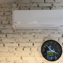 Termferm Klimatyzacje Pompy Ciepła Podhale - Biały klimatyzator VIVAX zamontowany na ścianie z białej cegły, obok czarny zegar ścienny wskazujący godzinę 10:20. Na tarczy zegara widoczne odbicie pokoju z zabawkami.
