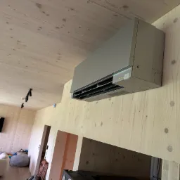 Mitsubishi klimatyzator ścienny zamontowany w nowoczesnym wnętrzu z boazerią, widoczne diody sygnalizacyjne i fragment salonu z sofą.