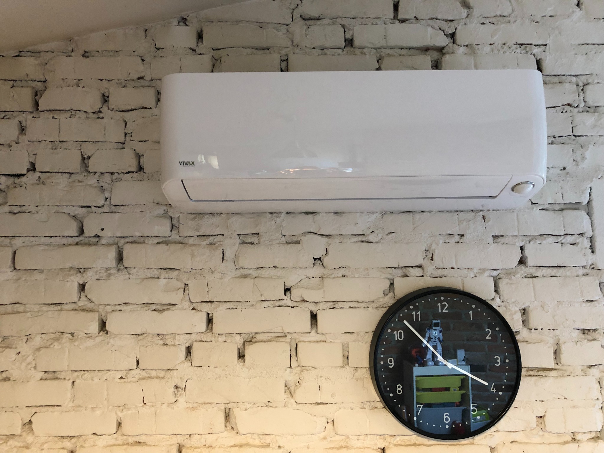 Biały klimatyzator VIVAX zamontowany na ścianie z białej cegły, obok czarny zegar ścienny wskazujący godzinę 10:20. Na tarczy zegara widoczne odbicie pokoju z zabawkami.