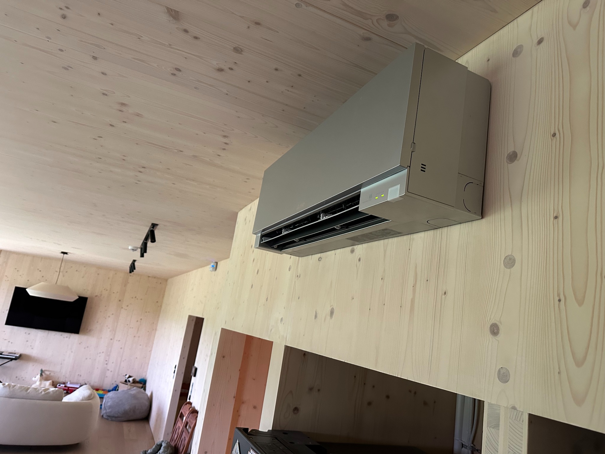 Mitsubishi klimatyzator ścienny zamontowany w nowoczesnym wnętrzu z boazerią, widoczne diody sygnalizacyjne i fragment salonu z sofą.