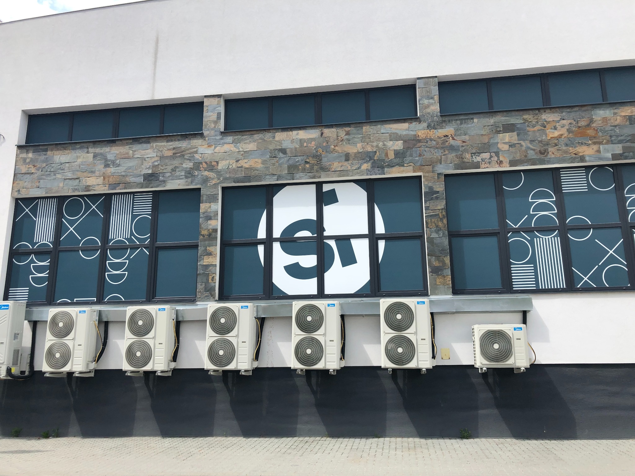 Rząd jednostek zewnętrznych klimatyzacji Midea zamontowanych na elewacji budynku z oknami ozdobionymi geometrycznymi wzorami i logo.