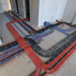 Równolegle ułożone wiązki przewodów elektrycznych i telekomunikacyjnych w rurkach karbowanych, mocowane metalowymi uchwytami do surowej, betonowej podłogi w nowym budynku, widok z góry.