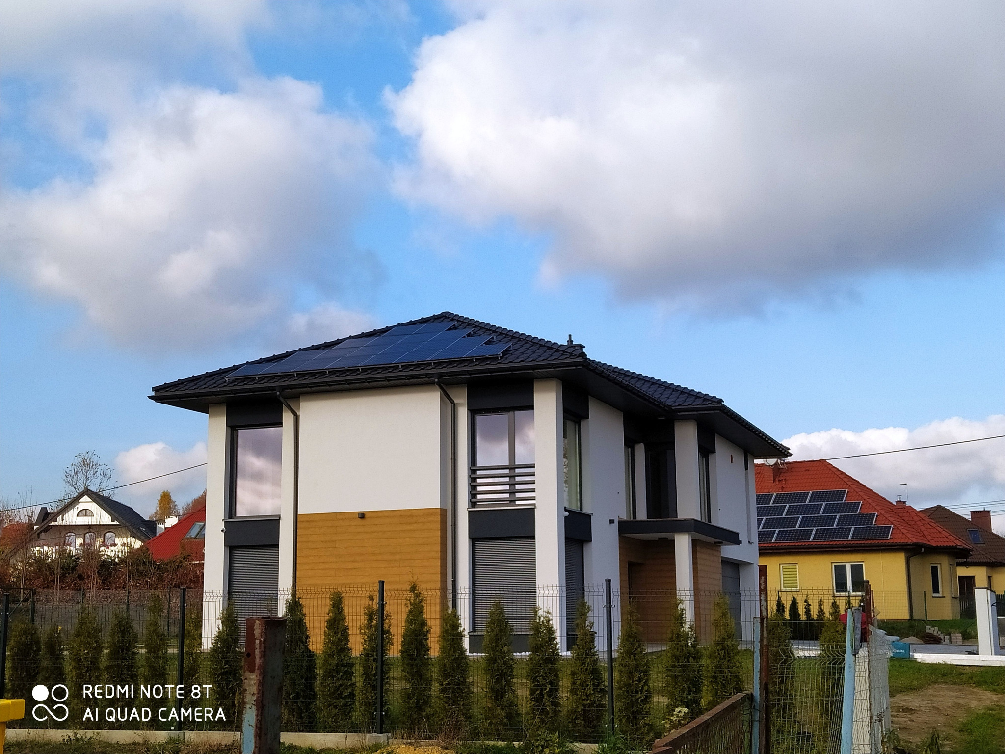 Nowoczesny dom jednorodzinny z panelami słonecznymi na dachu, widok zza ogrodzenia, w tle inne domy, również z panelami fotowoltaicznymi.
