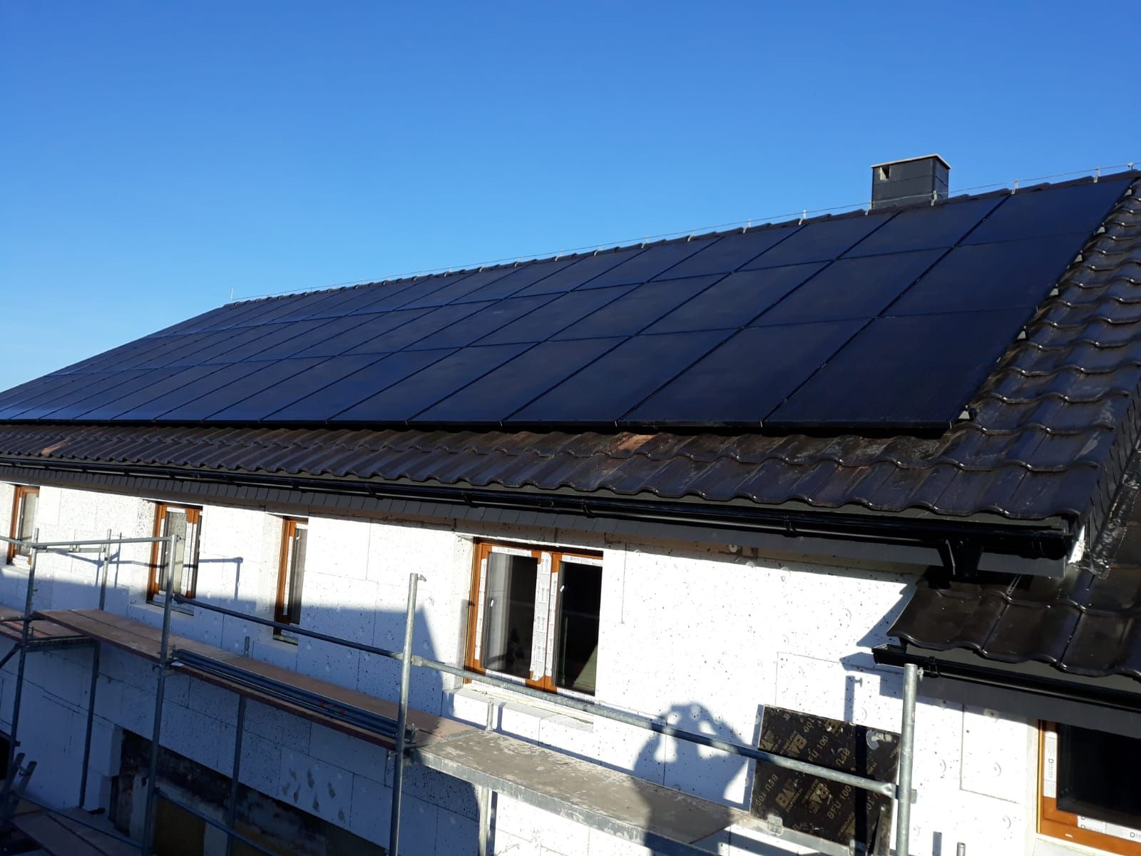 Panele fotowoltaiczne na dachu domu w trakcie budowy, widoczne rusztowania i niezakończona elewacja, błękitne niebo w tle.