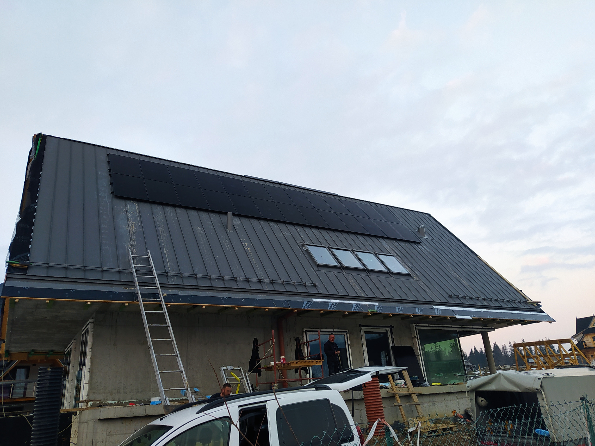 Dom w trakcie montażu paneli słonecznych na dachu z blachy, widoczna drabina i ekipa montażowa, w tle zalesiony teren.