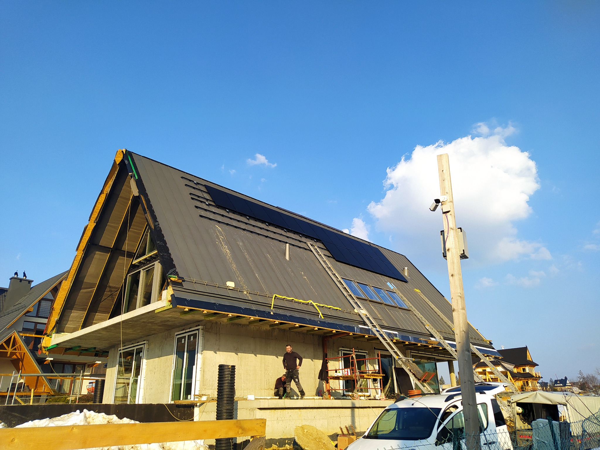 Montaż paneli na dachu domu w stylu góralskim, widoczny mężczyzna na rusztowaniu i drabina oparta o dach, błękitne niebo w tle.