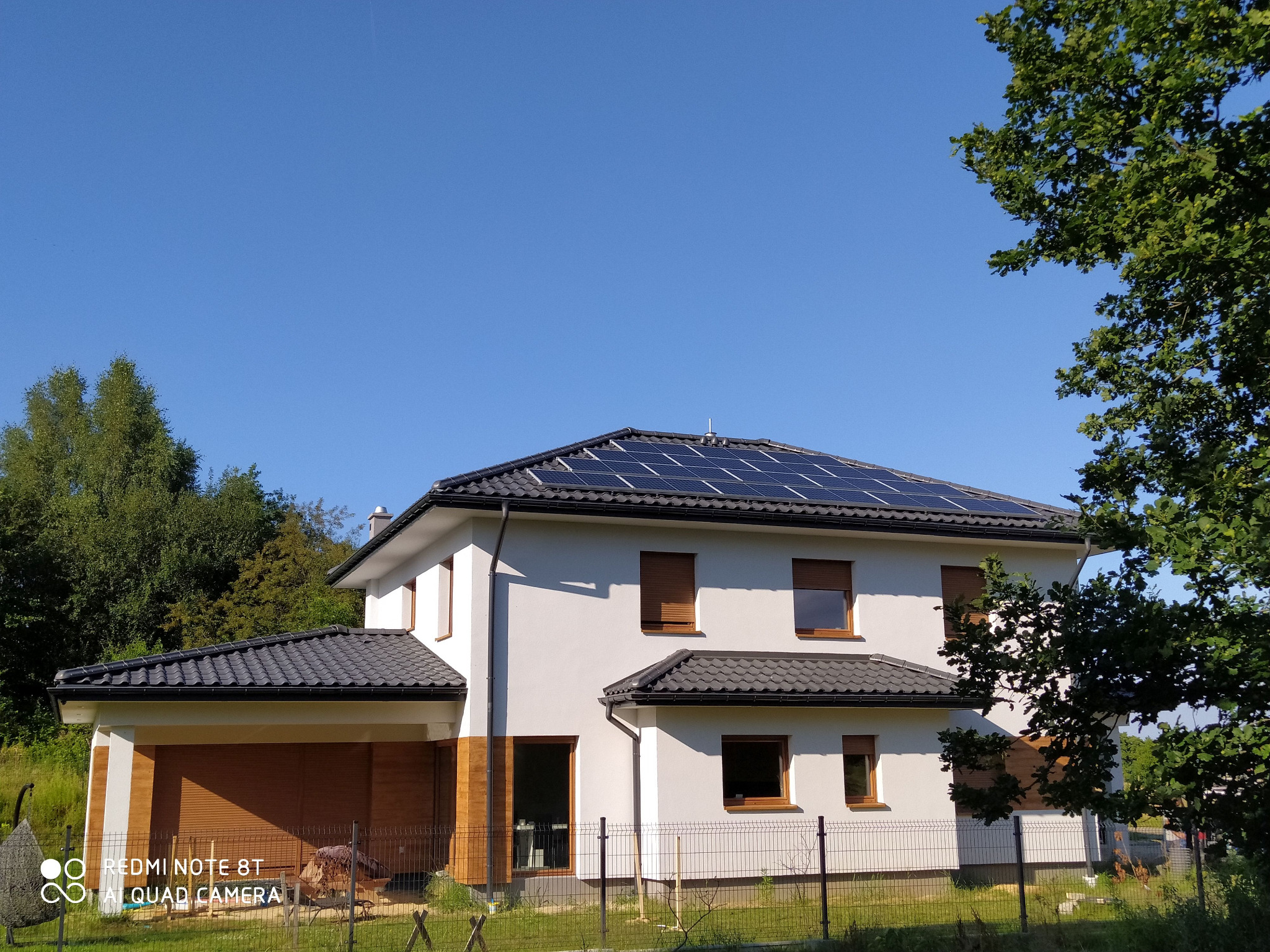 Dwukondygnacyjny dom jednorodzinny z szarym dachem i panelami słonecznymi, widziany w słoneczny dzień z perspektywy niskiego kąta, z zielenią drzew w tle.