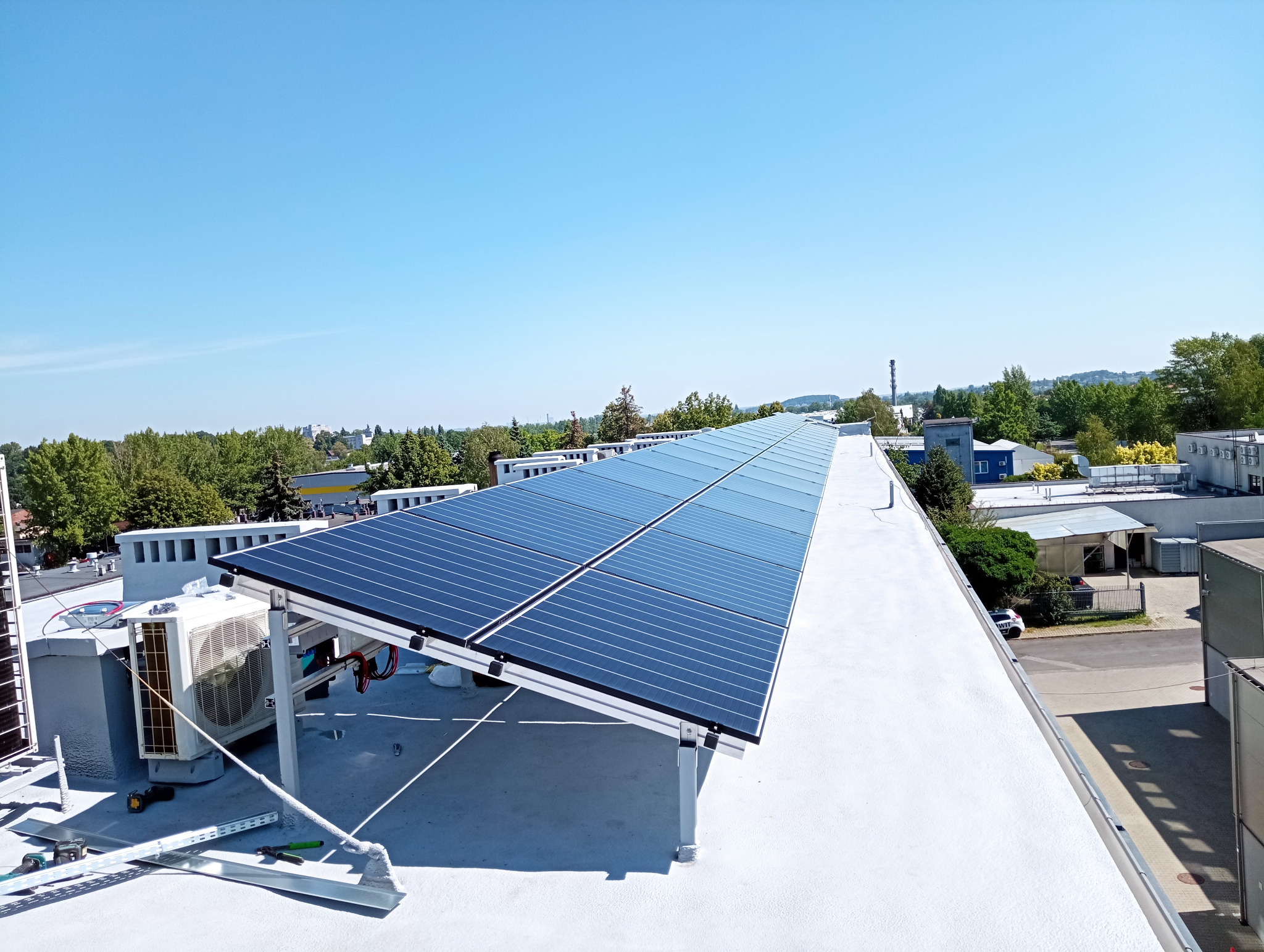 Instalacja fotowoltaiczna na płaskim dachu budynku przemysłowego, widoczne panele, inwerter i elementy montażowe, w tle zieleń i błękitne niebo.