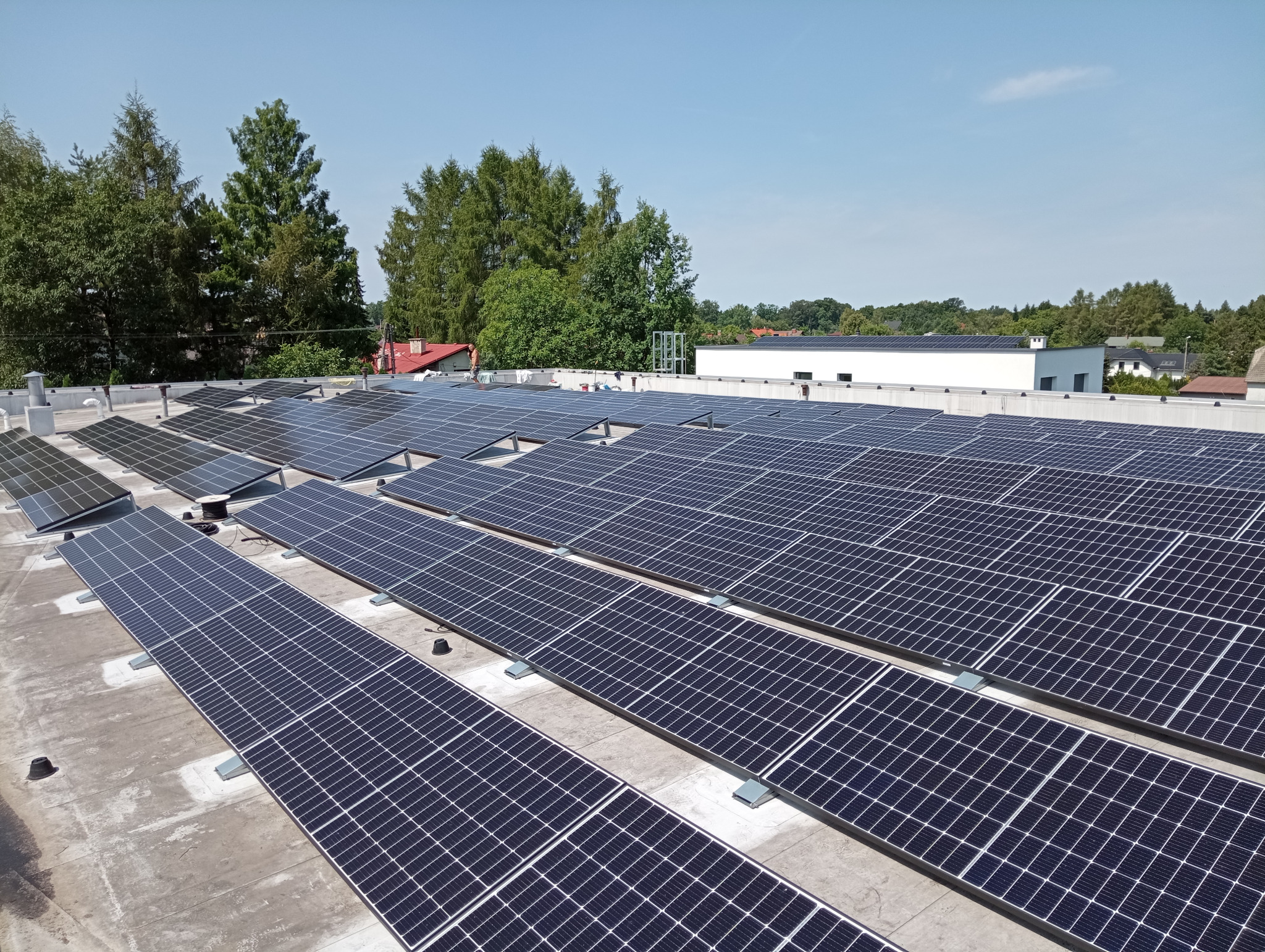 Rząd paneli słonecznych na dachu budynku, widok z góry, z zielenią w tle i błękitnym niebem.